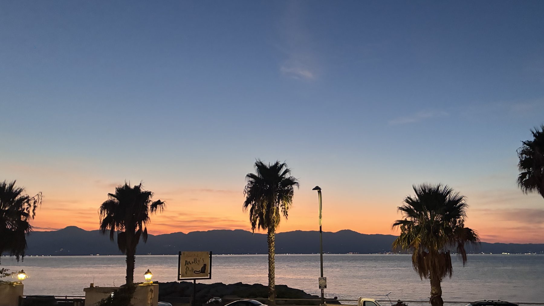 Il tramonto sullo Stretto di Messina visto dalla veranda del Molly's Pub: palme in controluce, l'insegna del pub illuminata e i monti della Sicilia all'orizzonte.